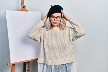 Beautiful caucasian woman standing by painter easel stand suffering from headache desperate and...
