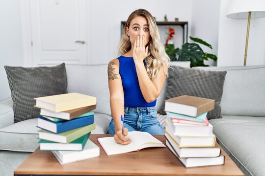 Young beautiful woman writing on notebook around university books covering mouth with hand, shocked and afraid for mistake. surprised expression