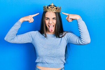 Young brunette teenager wearing princess crown sticking tongue out happy with funny expression.