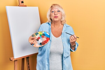 Middle age blonde woman standing drawing with palette by painter easel stand puffing cheeks with funny face. mouth inflated with air, catching air.