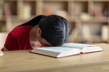 Boring tired unhappy upset teen asian girl lies on book in room or library interior