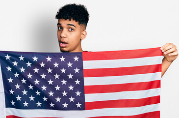 Young african american man holding united states flag in shock face, looking skeptical and...