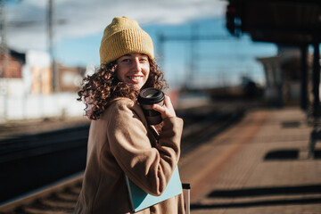 Portrait of happy young traveler woman with coffe and luggage at train station platform