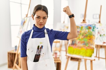 Young brunette woman at art studio strong person showing arm muscle, confident and proud of power