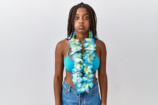 Young african american woman with braids wearing bikini and hawaiian lei puffing cheeks with funny face. mouth inflated with air, crazy expression.