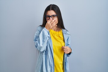 Young hispanic woman standing over blue background smelling something stinky and disgusting, intolerable smell, holding breath with fingers on nose. bad smell