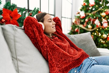 Young hispanic girl listening to music sitting on the sofa by christmas tree at home.