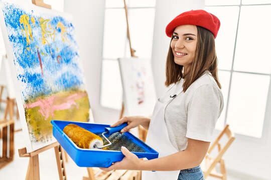 Young hispanic artist woman wearing french beret drawing using paint roller at art studio.