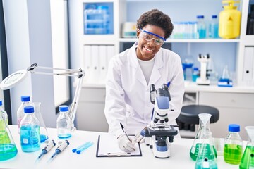 African american woman wearing scientist uniform using microscope write on clipboard at laboratory
