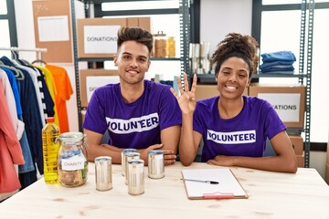 Young interracial people wearing volunteer t shirt at donations stand showing and pointing up with fingers number four while smiling confident and happy.