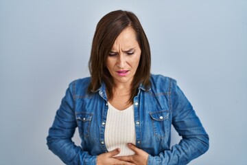 Middle age brunette woman wearing casual denim jacket over isolated background with hand on stomach...