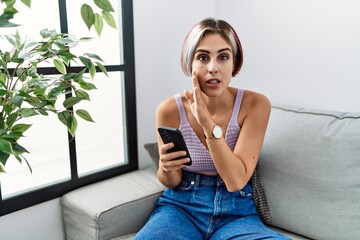 Young beautiful woman using smartphone typing message sitting on the sofa hand on mouth telling secret rumor, whispering malicious talk conversation