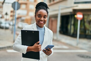 Young african american businesswoman using smartphone holding clipboard at the city.