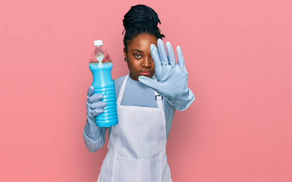Young african american woman wearing apron holding detergent bottle with open hand doing stop sign with serious and confident expression, defense gesture