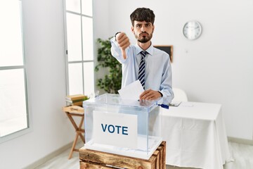 Hispanic man with beard voting putting envelop in ballot box looking unhappy and angry showing rejection and negative with thumbs down gesture. bad expression.