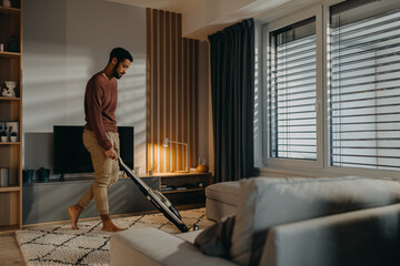 Young man vacuum cleaning carpet in living room - obrazy, fototapety, plakaty