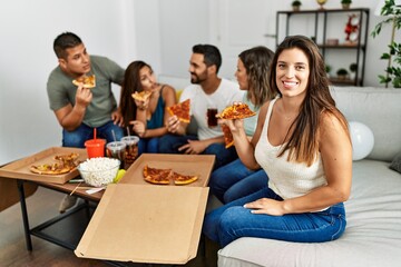 Group of young hispanic friends smiling happy eating italian pizza sitting on the sofa at home.