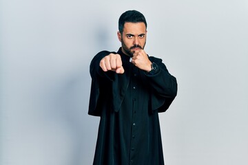 Handsome hispanic man with beard wearing catholic priest robe punching fist to fight, aggressive...