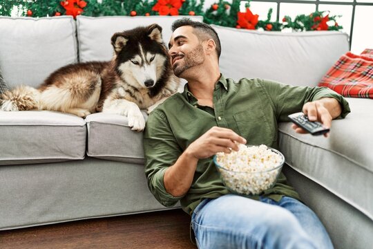 Young hispanic man watching movie sitting on sofa with dog by christmas decor at home