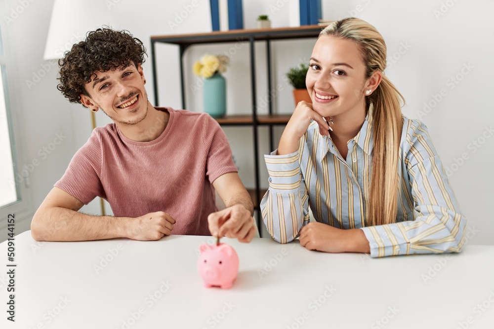 Poster Young couple inserting coin inside piggy bank at home.