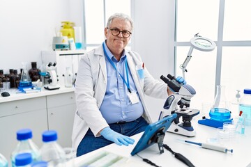 Middle age grey-haired man wearing scientist uniform using microscope and touchpad at laboratory