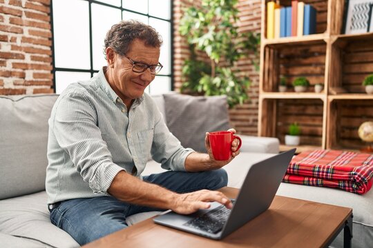 Middle age man using laptop and drinking coffee sitting on sofa at home