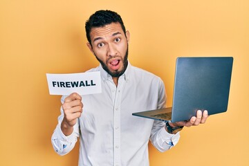 Hispanic man with beard holding computer laptop and firewall banner in shock face, looking...