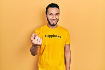 Hispanic man with beard wearing t shirt with happiness word message beckoning come here gesture with hand inviting welcoming happy and smiling