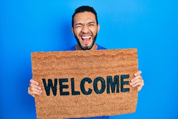 Hispanic man with beard holding welcome doormat smiling and laughing hard out loud because funny...