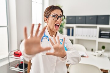 Young brunette doctor woman wearing stethoscope at the clinic afraid and terrified with fear expression stop gesture with hands, shouting in shock. panic concept.