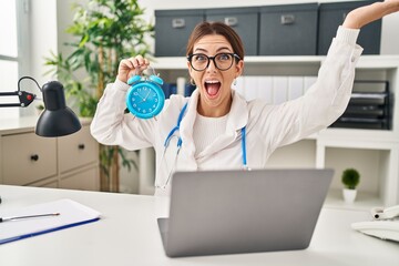 Young brunette doctor woman holding alarm clock celebrating victory with happy smile and winner expression with raised hands