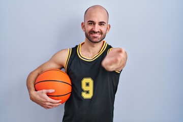 Young bald man with beard wearing basketball uniform holding ball smiling cheerful offering palm hand giving assistance and acceptance.