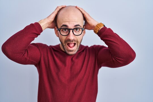 Young bald man with beard standing over white background wearing glasses crazy and scared with hands on head, afraid and surprised of shock with open mouth