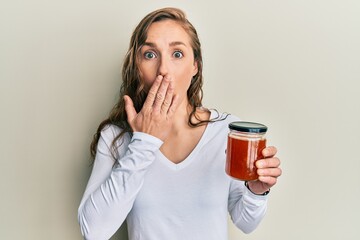 Young blonde woman holding jam covering mouth with hand, shocked and afraid for mistake. surprised...