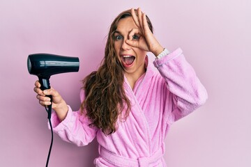 Young blonde woman wearing bathrobe using hair dryer smiling happy doing ok sign with hand on eye...