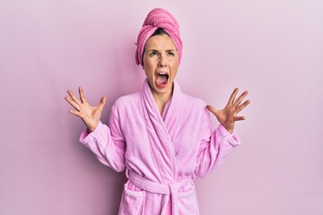 Young blonde woman wearing shower towel cap and bathrobe crazy and mad shouting and yelling with aggressive expression and arms raised. frustration concept.