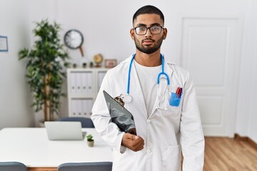 Young indian man wearing doctor uniform and stethoscope relaxed with serious expression on face....