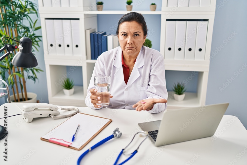 Poster hispanic mature doctor woman holding pill and glass of water depressed and worry for distress, cryin