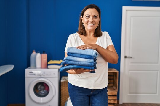 Middle age hispanic woman smiling confident holding folded jeans at laundry room