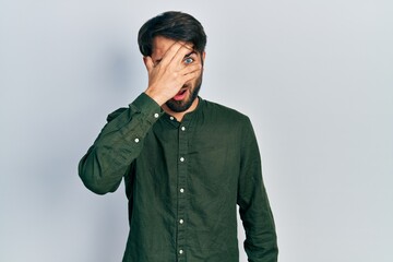Young hispanic man wearing casual clothes peeking in shock covering face and eyes with hand, looking through fingers afraid