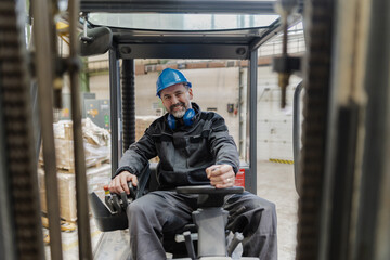 Happy mature man fork lift truck driver lifting pallet in storage warehouse and looking at camera.