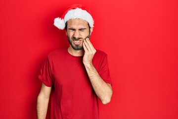 Young hispanic man wearing christmas hat touching mouth with hand with painful expression because of toothache or dental illness on teeth. dentist