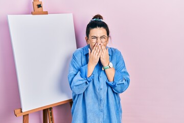 Young hispanic woman standing by painter easel stand laughing and embarrassed giggle covering mouth...