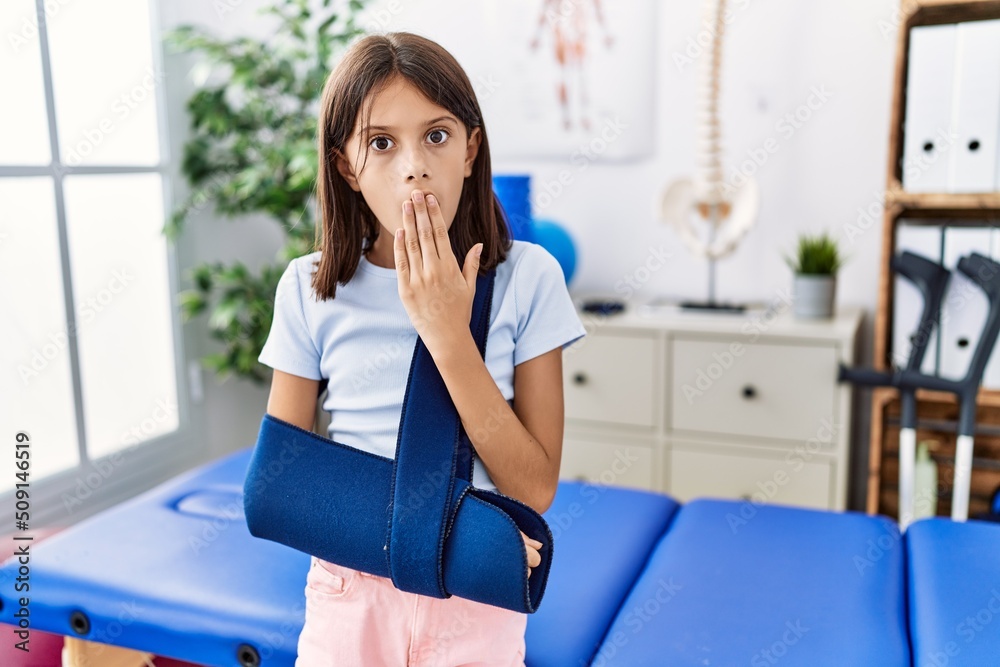 Poster Young hispanic girl wearing arm on sling at rehabilitation clinic covering mouth with hand, shocked and afraid for mistake. surprised expression