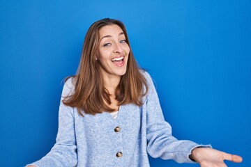 Young woman standing over blue background smiling cheerful with open arms as friendly welcome, positive and confident greetings