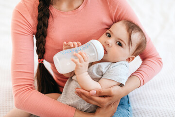 Closeup portrait of mother with baby on hands, little child holding bottle and drinking milk at home