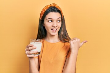 Young brunette girl drinking a glass of milk pointing thumb up to the side smiling happy with open...