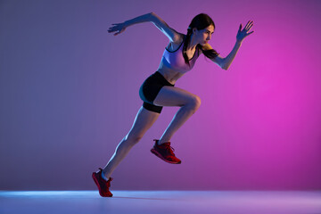 Obraz na płótnie Canvas One young muscular girl, female runner or jogger training isolated on pink-blue background in neon light. Sport, track-and-field athletics, competition and active lifestyle concept