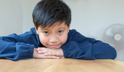 Obraz premium Cute Asian boy with white skin smiling looking in camera happily resting chins on hands-on wooden table. Close-up portrait of a cute child.