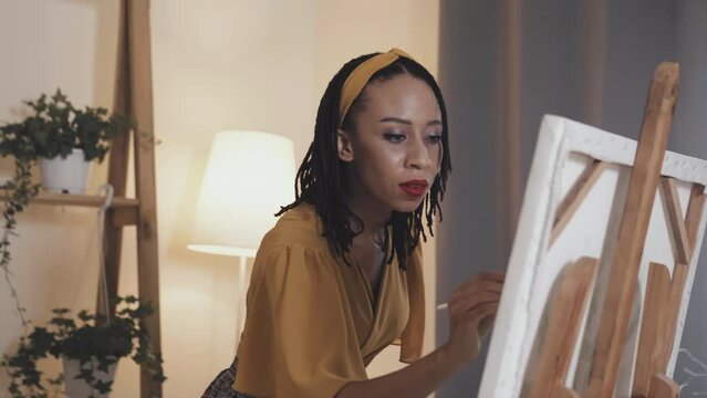 Beautiful African American woman painting picture on canvas with brush and looking at it while spending leisure time at home